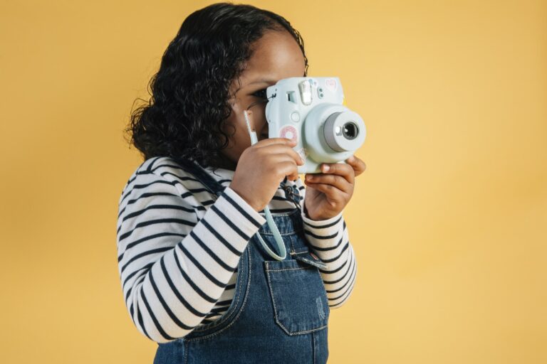 Focused little African American girl in casual wear taking pictures on contemporary instant photo camera while standing against yellow background