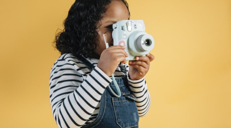Focused little African American girl in casual wear taking pictures on contemporary instant photo camera while standing against yellow background