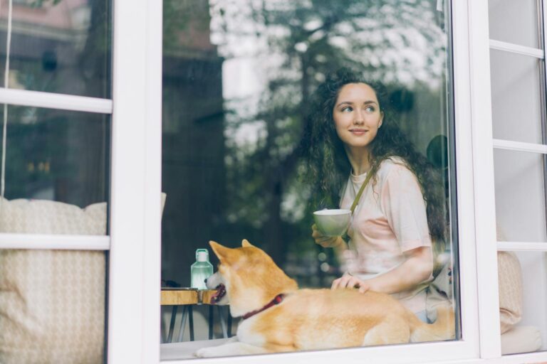 Relaxed woman enjoying a warm beverage with a Shiba Inu dog by a cozy window view.